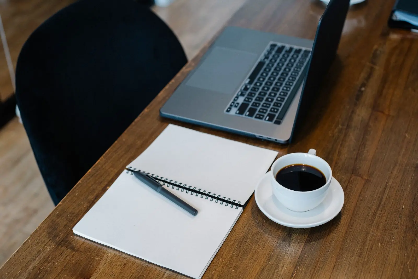 A laptop, an open notebook and a cup of black coffee on a wooden desk